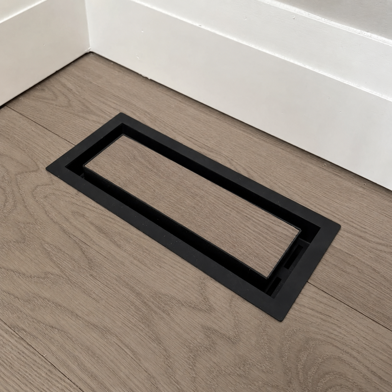 Black floor-matching vent cover installed in wood flooring near a white wall
